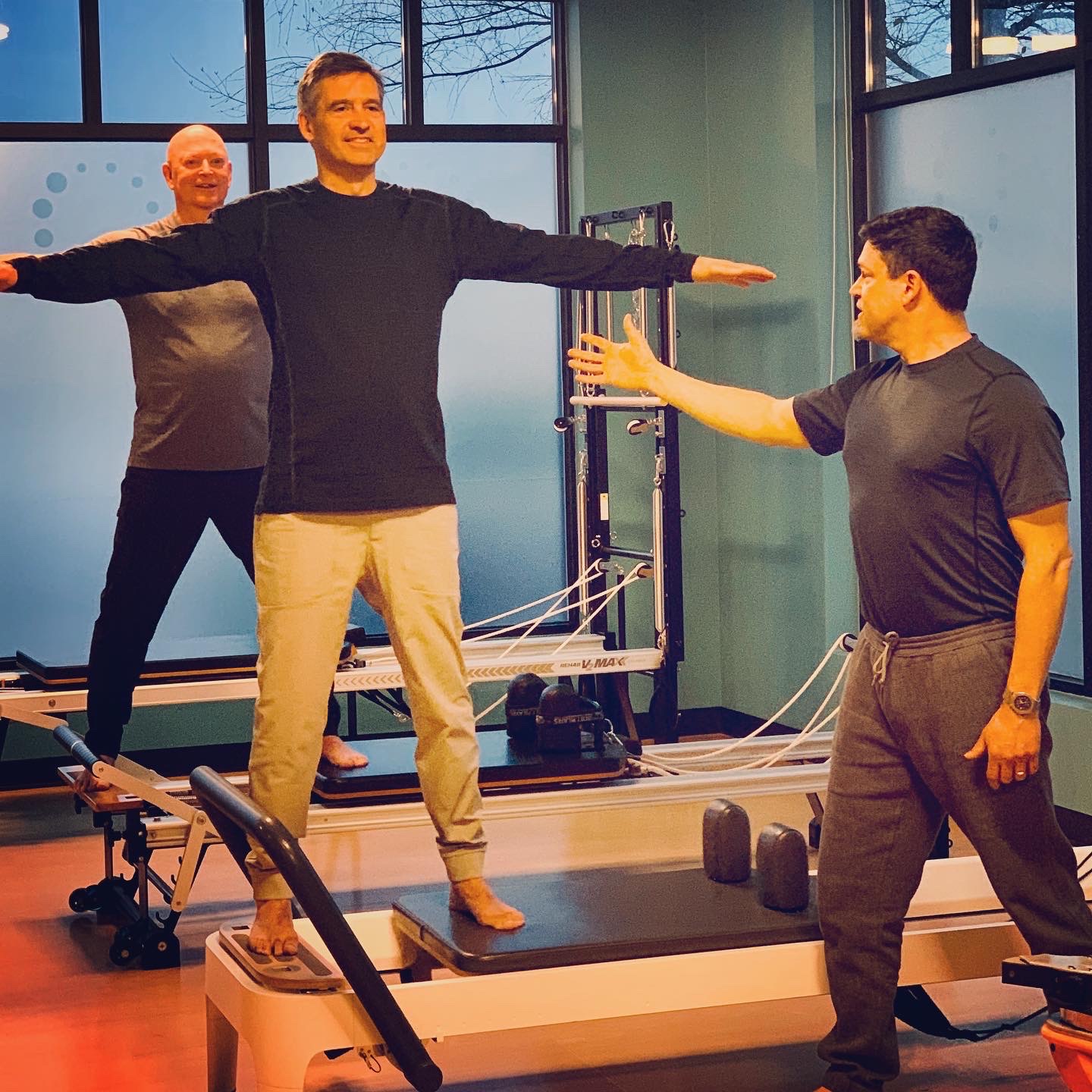 Two men working out with a trainer on the Pilates Reformer